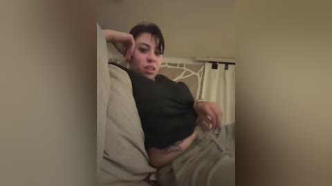 Video of a woman with medium skin tone, short dark hair, wearing a black top and beige pants, lying on a beige bed, looking tired. Background shows beige walls and a white curtain.