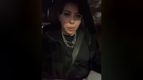Video of a young woman with dark hair, wearing a black jacket, seated in a car, dim lighting, chain necklace, blurred background.