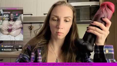 A video of a young woman with light skin and long brown hair, wearing a plaid shirt, holding a large, realistic-looking pink dildo, set in a modern kitchen.