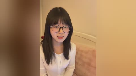 Video of a young East Asian woman with straight black hair, wearing glasses and a white top, seated on a plush pink couch against a beige wall.