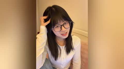 Video of an Asian woman with straight black hair, glasses, and a white long-sleeved top, smiling, adjusting her hair, standing in a beige room.