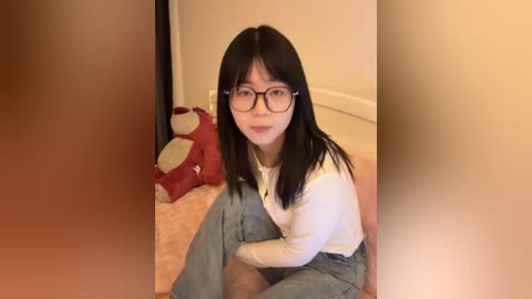 Video of an Asian woman with straight black hair, wearing glasses, a white shirt, and blue pants, sitting on a pink carpet in a beige room with a red stuffed animal.