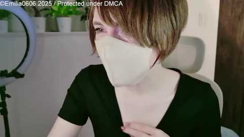 Video of a young woman with short, blonde hair wearing a black top and white mask, sitting indoors by a ring light, captioned \"EmiliaOgO 2023/04/23, Produced under DMCA.\