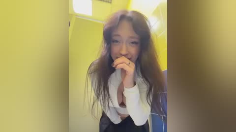 A video of a young Asian woman with long brown hair, wearing a white long-sleeve top and black skirt, posing in a yellow bathroom, smiling and covering her mouth with her fingers.