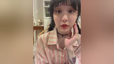 A video of a young Asian woman with pale skin, wearing a pink and white striped blouse, a black choker, and a tattoo on her right wrist. She has long black hair and is sitting indoors with a neutral expression.