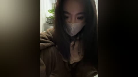 Video of an East Asian woman with long black hair, wearing a beige face mask and a brown coat, peering through a dark doorway into a dimly lit room with green potted plants on a shelf.