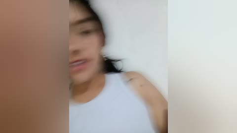 A blurred video of a woman with light skin and dark hair, wearing a white tank top, lying on a white pillow. The image is out of focus, making details indistinct.