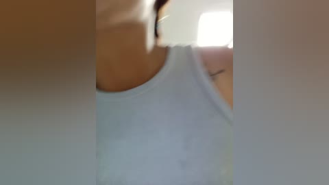 Blurry video of a woman in a white tank top, with a slight focus on her bare shoulder, in a dimly lit room with a window visible in the background.
