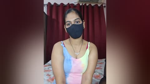 Video of a woman with medium brown skin and dark hair in a ponytail, wearing a black face mask, a tie-dye tank top, and a necklace, sitting on a bed with red curtains in the background.