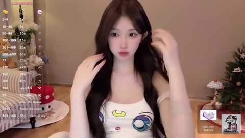 Video of an East Asian woman with long black hair, fair skin, and a slender physique, wearing a white tank top with cartoon glasses, sitting in a cozy, festive living room with a Christmas tree and decorations.