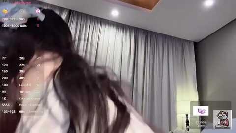 Video of a woman with long, dark hair, blurred, in a room with gray curtains and a lamp. Text overlays show viewer stats and a small chat window.