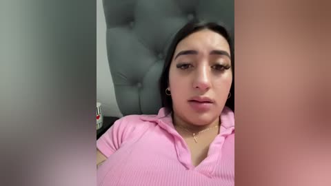 Media: Video of a young woman with medium skin tone, dark hair, and wearing a pink top, lying on a tufted gray couch in a dimly lit room.