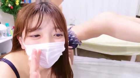 A close-up video of an Asian woman with light skin, brown hair, wearing a white face mask, and blue-patterned bra, giving a hand job to a partially visible person on a massage table in a dimly lit room.