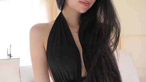 Video of an Asian woman with long black hair, wearing a black halter dress, in a softly lit room with white curtains.