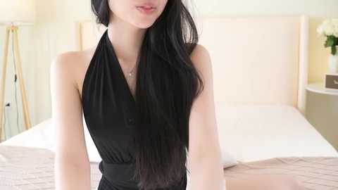 Video of a slim, fair-skinned woman with long, straight black hair, wearing a black halter dress, standing in a bright, minimalist bedroom with a white bed and beige walls.