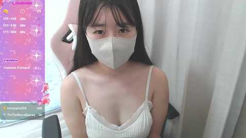 Video of an East Asian woman with long black hair, wearing a white mask and white camisole, sitting in a room with a pink digital screen displaying camera settings.
