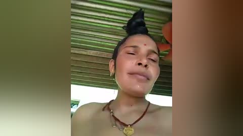 A close-up video of a smiling young woman with a high bun, wearing a brown beaded necklace, under a corrugated metal roof.