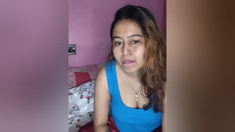 Video of a young woman with light brown skin and long, wavy brown hair, wearing a bright blue tank top, seated on a bed with red and white floral sheets, in a pink-painted room.