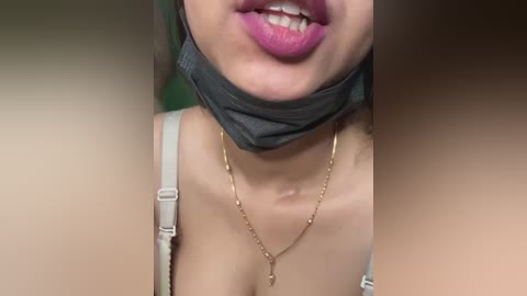 Close-up video of a woman with light skin, wearing a grey face mask, silver bra straps, and a gold necklace, smiling with visible teeth.