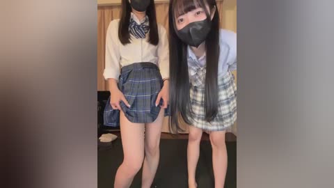 Video of two East Asian women in school uniforms, one with long black hair, wearing a black face mask, bending forward, the other with short black hair, standing straight. Background shows a wooden door and a green floor.