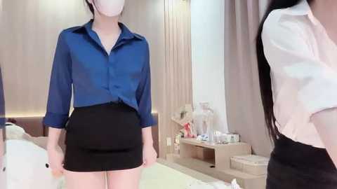 Video of a young Asian woman in a blue button-up shirt and black skirt, standing in a modern, sparsely furnished bedroom with light wood accents and a bed in the background.