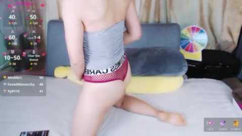 Video of a fair-skinned person in gray shorts with \"CAVA\" printed on the waistband, kneeling on a bed with yellow and gray pillows, in a bedroom with a colorful, circular wall decoration.