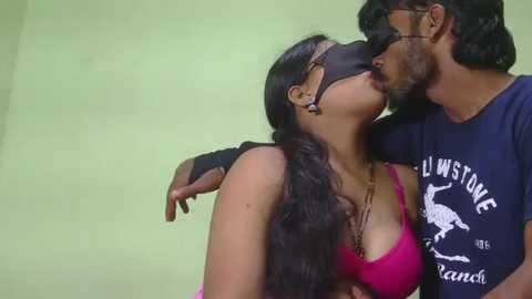 A video of a dark-haired woman in a pink bra and black blindfold kissing a bearded man in a dark T-shirt, both in a dimly lit room.