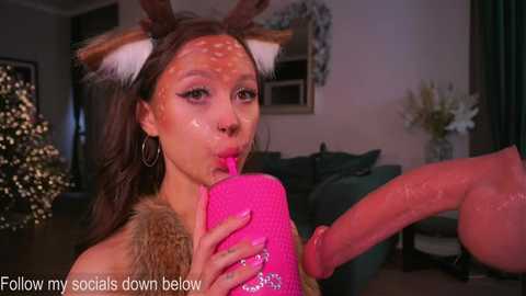 A photo of a young Asian woman with deer antler headpiece, wearing a fur-lined coat, drinking from a pink dildo, in a festive, dimly lit living room with Christmas decorations.