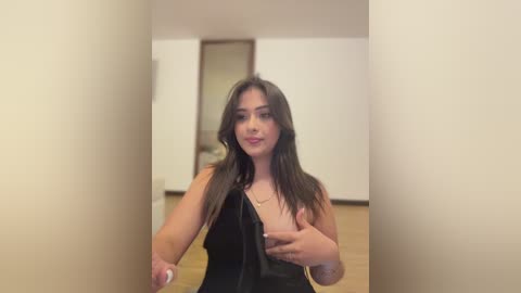 Video of a young, light-skinned woman with long, dark hair, wearing a black dress, standing in a minimalist room with beige walls and wooden floors, holding a phone.
