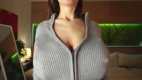 Video of a woman in a tight, ribbed, light grey zip-up sweater, highlighting her ample cleavage. Background features a modern living room with greenery outside the window and a potted plant on the left.