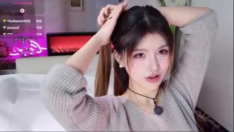Video of an Asian woman with long, dark hair, styled in a ponytail, wearing a gray knit sweater. She has a fair complexion, light makeup, and is indoors with a modern, minimalist decor.