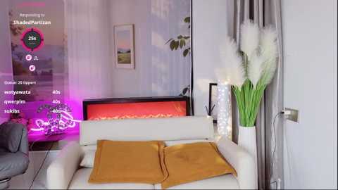Video of a modern living room with a white sofa adorned with two mustard-yellow cushions, a white vase with white feathers, a glass table, and a glowing fireplace in the background.