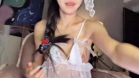 Media: Video of a young woman with pale skin and long black hair, wearing a sheer white lace bra, with a decorative feather and flowers on the left. Background shows a dimly lit room with a wall hanging.
