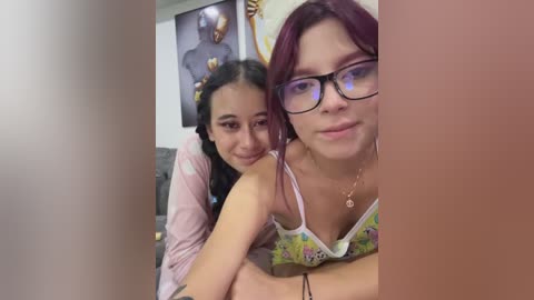 Video of two young women with light skin and long dark hair, one with glasses, lying close together on a bed. Background shows framed art and a desk.