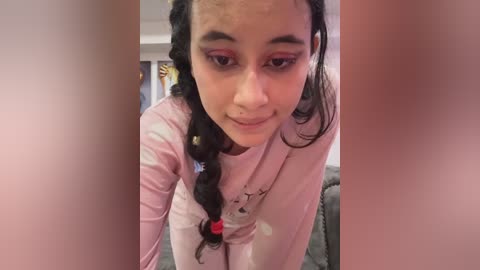 A video of a young girl with light brown skin and braided black hair adorned with a red hair tie. She wears pink eyeshadow and a light pink shirt. The background shows a living room with a couch and decorative items.