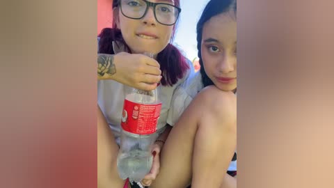 Video of two women with fair skin, one with red hair and glasses, holding a red cup, and the other with dark hair, sitting on a bed.