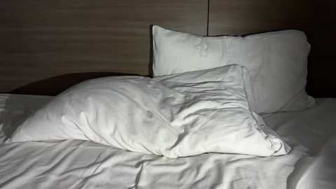 Video of a bed with a rumpled white duvet and two white pillows, set against a dark wooden headboard. The room is dimly lit, creating shadows and emphasizing the soft textures.