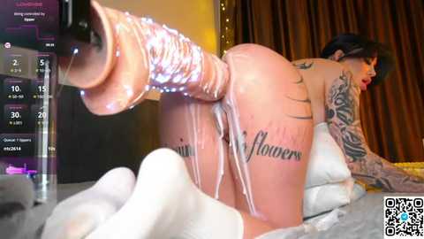 Video of a tattooed, fair-skinned woman with short dark hair, wearing white socks and a large, realistic flesh-colored dildo, positioned on all fours on a bed with a digital clock and a QR code in the background.