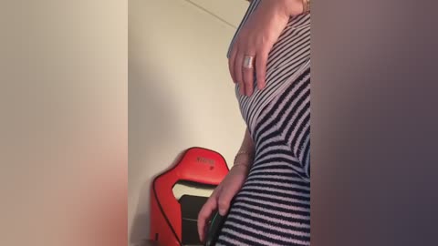 Media: Video of a pregnant person in a striped dress, hand resting on their belly, wearing a silver ring. Background features a red exercise ball and beige wall.