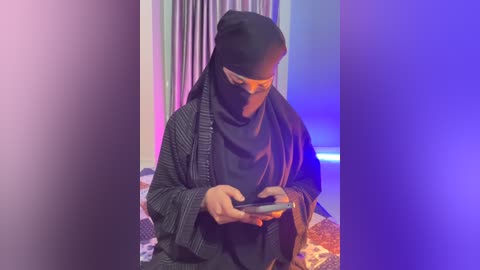 Video of a person in a black hijab and striped shawl, holding a smartphone, standing in a room with vertical, striped curtains and blue-purple lighting.