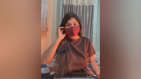 Video of a young South Asian woman with long, dark hair, wearing a maroon face mask and a dark T-shirt, sitting on a bed with patterned blankets. She adjusts her mask with her right hand. The background features a wall with a lace curtain and a wooden door.