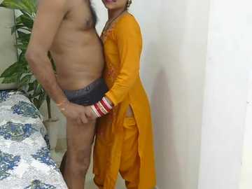 Media: Video of a shirtless man in underwear, holding a woman in an orange salwar kameez, standing indoors near a white wall and patterned bedspread.