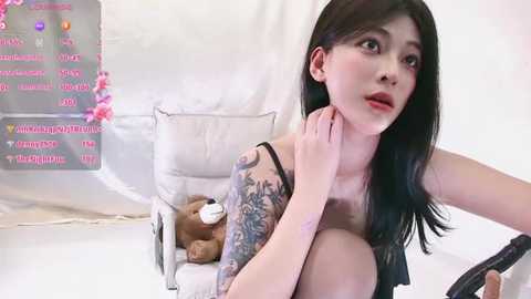Video of a young East Asian woman with long black hair, wearing a black tank top, sitting on a white chair with a teddy bear, in a minimalist, white room.