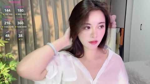 Video of a young Asian woman with fair skin and long brown hair, wearing a white blouse, posing indoors with a plant in the background.
