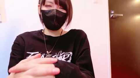 Media: Video of a young Asian woman with short black hair, wearing a black face mask and a black sweatshirt with white text, holding a small dog in her arms. Background features a white wall and a dark wooden door.