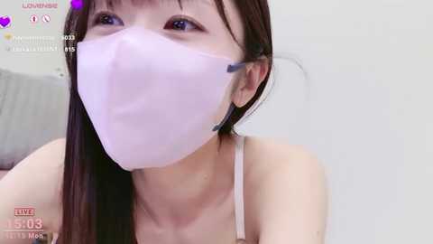 A video of a young Asian woman with long black hair, wearing a light pink face mask and headphones, sitting in a white room.