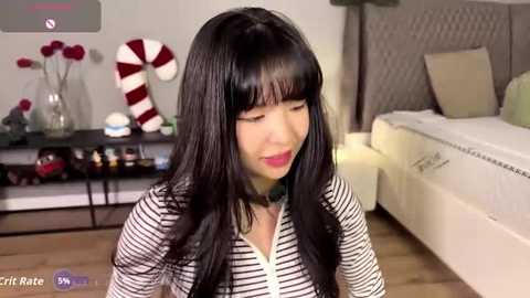 A video of a young Asian woman with long black hair, wearing a striped shirt, sitting on a wooden floor in a modern bedroom with a white bed, a wall-mounted shelf, and red-and-white candy canes.