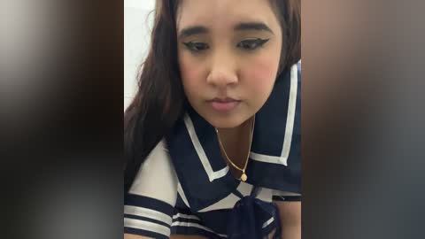 A video of an Asian woman with long, dark hair, wearing a navy blue and white striped polo shirt, with black eyeliner and a delicate gold necklace, looking down with a thoughtful expression.
