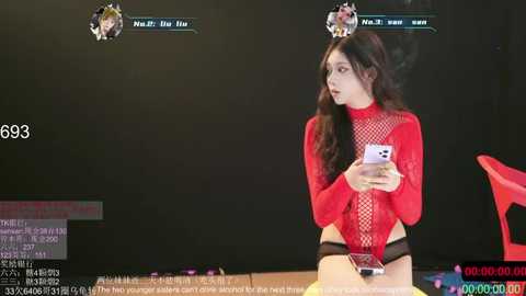 A video shows an East Asian woman with long, wavy black hair in a revealing red fishnet top and black panties, seated on a table, holding a smartphone. The background features a black curtain and a red chair.