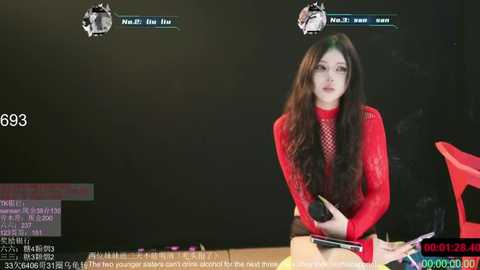 Video of a woman with long black hair, fair skin, and a slender figure, wearing a red lace top, sitting at a desk. She holds a phone and laptop. Background shows a dark room with two virtual avatars above her.
